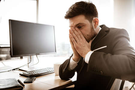 Business crisis concept. Emotional young businessman at work. Worried stressed manager sitting at the office table grabs his face with hands. Busy man in suit indoors shows negative emotions.の写真素材