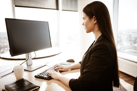 Business concept. Successful young businesswoman at work. Manager sitting at the office table and working on computer. Busy woman in suit indoors on glass window backgroundの写真素材