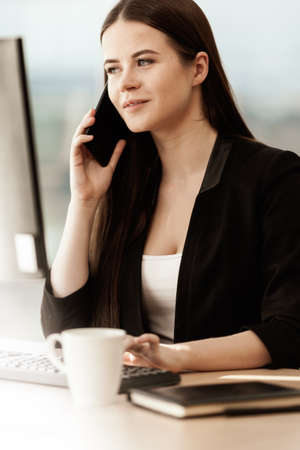 Business concept. Young businesswoman sitting at the office table happy talking on a cell phone getting good news about her work. Hands on keyboard. Woman in suit indoors on glass window backgroundの写真素材