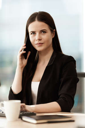 Business concept. Young businesswoman sitting at the office table happy talking on a cell phone getting good news about her work. Hands on keyboard. Woman in suit indoors on glass window backgroundの写真素材