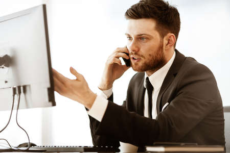 Business crisis concept. Young businessman sitting at the office table busy talking on a cell phone resolving a very serious work problem. Man in suit indoors on glass window backgroundの写真素材