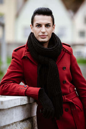 LGBTQ community lifestyle concept. Young homosexual man stands near stone fence. Handsome fashionable gay male model poses in cityscape outdoors. Wears red coat, gloves, and black scarfの写真素材