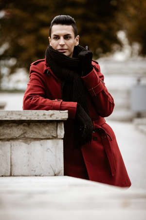 LGBTQ community lifestyle concept. Young homosexual man stands near stone fence. Handsome fashionable gay male model poses in cityscape outdoors. Wears red coat, gloves, and black scarfの写真素材