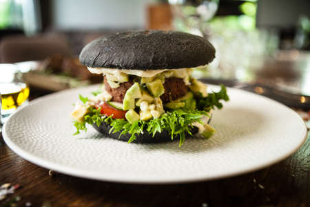 Vegan burger. Black bun, tofu-avocado salad, mayonnaise, cucumber, tomato, fresh salad. Delicious healthy hamburger food closeup served on a table for lunch in modern cuisine gourmet restaurant.の写真素材