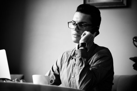 LGBT community lifestyle concept. Young homosexual man sits at the table in old-fashioned city cafe. Handsome gay male businessman talks on a phone having a break at lunch. Black and white monochrome.の写真素材