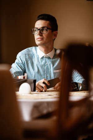 LGBTQ community lifestyle concept. Young homosexual man sits at the table in old-fashioned city cafe. Handsome smiling gay male businessman poses while having a break at lunchの写真素材