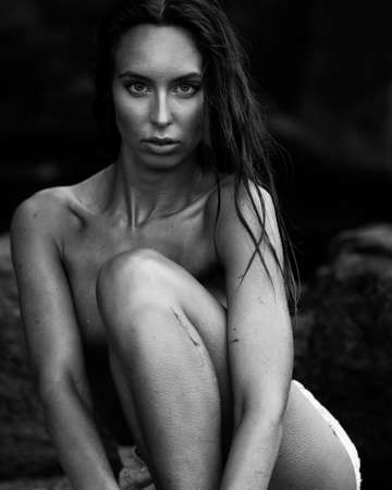 Summertime recreation concept. Beautiful young sexy woman with fit trained slim body wearing blue shorts sits on a beach on a stone. Fashion female model poses by the sea. Black and white monochrome.の写真素材