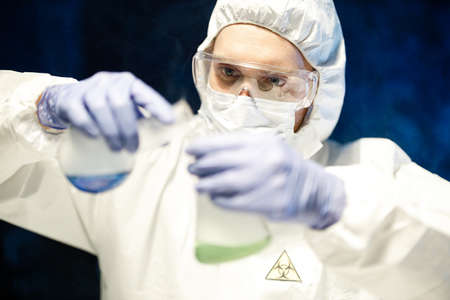 Corona virus concept. Male scientist doctor in mask, glasses and protective suit making biochemical experiment using glass flasks with colorful liquids in laboratory during COVID pandemic outbreakの写真素材