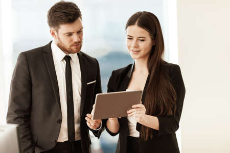 Successful business and modern technology concept. Young businesswoman and happy smiling businessman working together using tablet computer. Two colleagues man and woman teamwork indoors.の写真素材