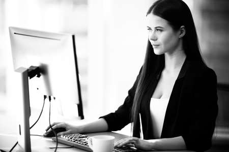 Business concept. Successful young businesswoman at work. Manager sits at the office table and works on computer. Busy woman in suit indoors on glass window background. Black and white, monochrome.の写真素材