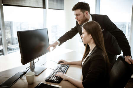 Business crisis concept. Young businesswoman sits at the office table busy working on computer. Businessman points at monitor. Resolving a serious work problem. Two managers man and woman indoors.の写真素材