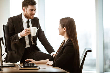 Flirt and relations at work. Smiling male businessman sits and shows sexual interest to positive minded female assistant at workplace. Two people man and woman love and sympathy in modern officeの写真素材