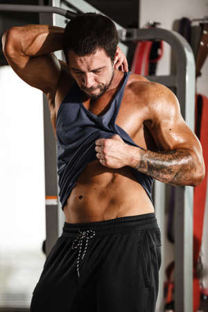 Fitness in gym, sport and healthy lifestyle concept. Handsome athletic man in blue shirt demonstrates his abs. Bodybuilder male model showing trained body.の写真素材