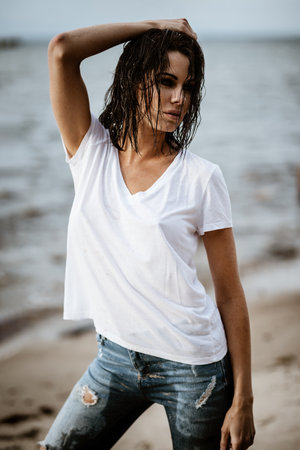 Summertime recreation concept. Beautiful young sexy woman with fit trained slim body wearing blue jeans and white t-shirt stands on a beach. Fashion female model poses by the sea.の写真素材