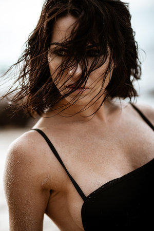 Summertime recreation concept. Beautiful young sexy woman with fit trained slim body wearing black swimwear bikini sits on a beach. Fashion female model poses by the sea.の写真素材
