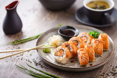 Baked sushi maki rolls with salmon, crab and spicy sauce on a plate with chopsticks, soy sauce, wasabi and ginger. Japanese traditional fish food closeup served for lunch in modern gourmet restaurant.の写真素材