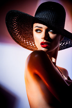 Fashion concept. Portrait of a beautiful woman in big black hat in bright mixed contrast light. Female model wearing accessories poses in studio. Professional makeup and colorful background.の写真素材