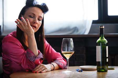 Alcoholism problem concept. Sad middle aged woman in stress sits in the kitchen, drinks white wine from a glass and smokes a cigarette. A depressed female is addicted to alcohol at homeの写真素材