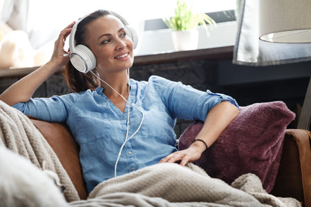 Leisure time concept. Happy beautiful woman listening to the music using headphones sitting on a couch indoors. Female spending her free day and relaxing at home aloneの写真素材
