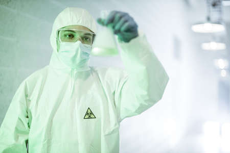 Male scientist doctor in mask, glasses and protective suit reviewing test-tube with blue liquid during pandemic outbreak. Man in biochemistry laboratory.の写真素材