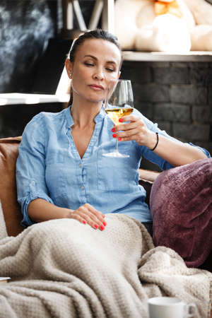 Leisure time concept. Happy beautiful woman drinks white wine from glass sitting on a couch indoors. Female spending her free day and relaxing at home alone.の写真素材