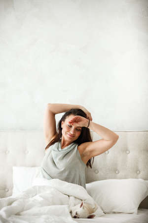 Lazy morning concept. Beautiful happy woman wakes up in bed and streches hands. Attractive female resting at home in bedroom. Little dog sleeps near on white sheets.の写真素材
