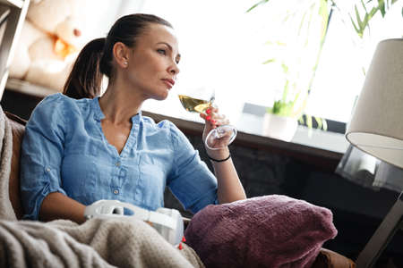 Leisure time concept. Happy beautiful woman drinks white wine from glass sitting on a couch indoors. Female spending her free day and relaxing at home alone.の写真素材