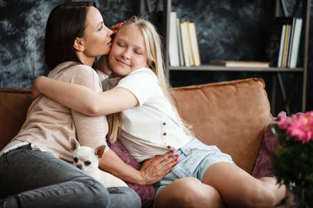Mother and daughter relationship concept. A woman and a teenage girl are sitting in front of the TV on the couch and have fun talking. The family spends time at home. Little dog lying on lapsの写真素材