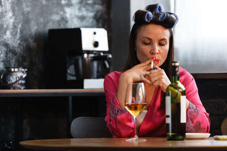 Alcoholism problem concept. Sad middle aged woman in stress sits in the kitchen, drinks white wine from a glass and smokes a cigarette. A depressed female is addicted to alcohol at home.の写真素材