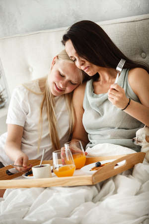Having family breakfast in bed concept. Young happy mother eating pancakes and drinking orange juice with her teenage daughter. Woman and girl sitting in the bedroom in the morning.の写真素材