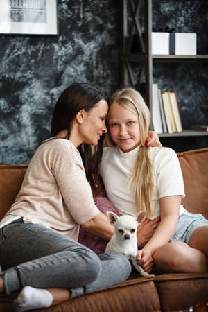 Mother and daughter relationship concept. A woman and a teenage girl are sitting in front of the TV on the couch and have fun talking. The family spends time at home. Little dog lying on lapsの写真素材