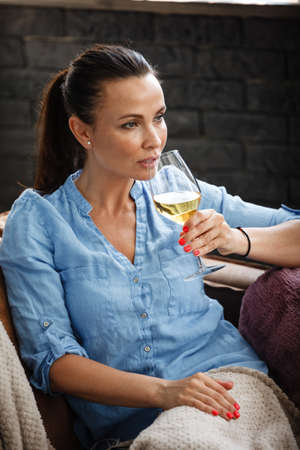 Leisure time concept. Happy beautiful woman drinks white wine from glass sitting on a couch indoors. Female spending her free day and relaxing at home alone.の写真素材