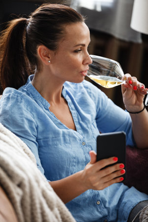Leisure time concept. Happy beautiful woman talks on a phone and drinks white wine from glass sitting on a couch indoors. Female spending her free day and relaxing at home alone.の写真素材