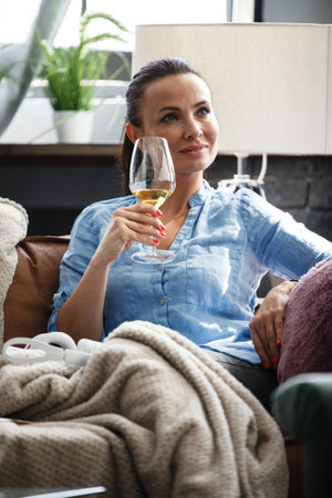 Leisure time concept. Happy beautiful woman drinks white wine from glass sitting on a couch indoors. Female spending her free day and relaxing at home alone.の写真素材