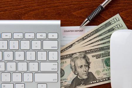 online banking  keyboard and mouse frame deposit slip and twenty dollar bills  pen points to deposit slip all are on top of a brown grain woodの写真素材