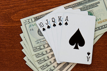 Spade royal flush sits on top of a stack of twenty dollar bills  brown grain wooden desktop...