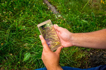 The young man holding mobile phone in his hands while sitting on the grass and taking some photos