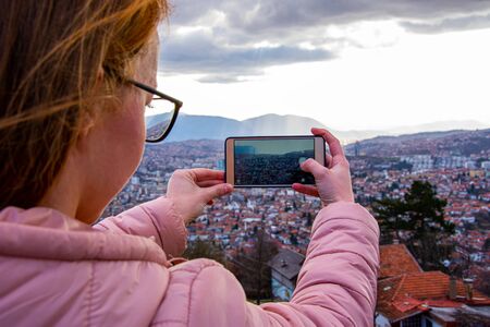 Girl with glasess and brown hair photographing cityscape of Sarajevo capital of BiH with mobile phoneの写真素材