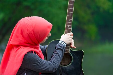Muslim girl wearing hijab holding guitar and looking to it in the nature during hazeの写真素材