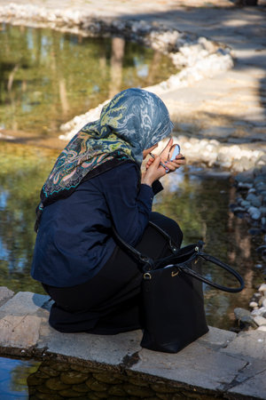 Muslim girl putting makeup in the park. Female model holding mirror and putting lipstic while she is...