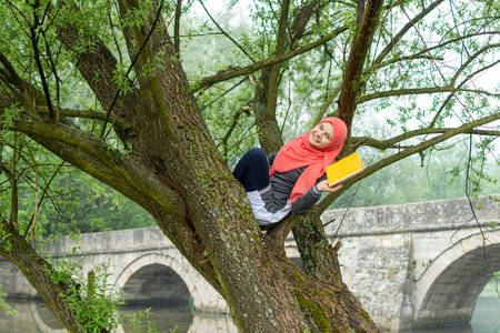Muslim girl smiling and holding the book while sitting on tree branch