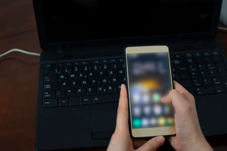 Woman's hand holding mobile phone with the laptop in the background. Woman trying to call laptop support or service for broken technical device. Lifestyleの写真素材