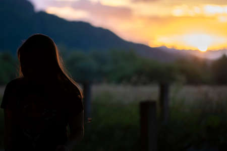 Silhouette of young teen girl sitting alone on the bench and beautiful landscape background behind her. Beautiful sunset time in the natureの写真素材