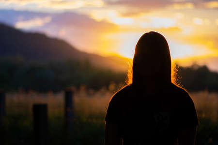 Silhouette of young teen girl sitting alone on the bench and beautiful landscape background behind her. Beautiful sunset time in the natureの写真素材