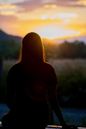 Silhouette of young teen girl sitting alone on the bench and beautiful landscape background behind her. Beautiful sunset time in the natureの写真素材
