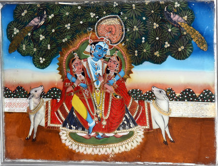 The image depicts a traditional Hindu painting of Lord Krishna with Radha and Rukmini, set within an ornate frame. Krishna, the central figure, is depicted with blue skin, adorned with jewelry and a crown, seated with his consorts. The background features a lush, stylized landscape with peacocks and floral motifs, characteristic of devotional art.の写真素材