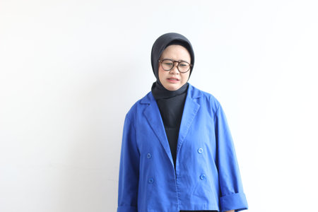 Beautiful young Asian Muslim woman, wearing glasses and blue blazer with disgusted face expression...