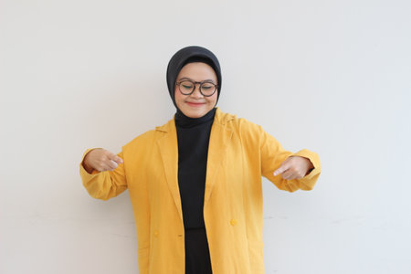 Beautiful young Asian Muslim woman in glasses, hijab and wearing yellow blazer smiling confident and happy with both hands pointing down isolated on white background, advertising conceptの写真素材