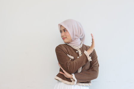 A young Muslim woman in a stylish brown jacket and light beige hijab poses against a plain white backdrop. The image is a studio portrait highlighting the fashion and style of the attire.の写真素材