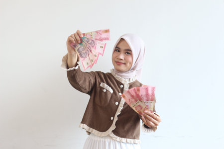 A young woman in a hijab holds Indonesian Rupiah banknotes.  She is smiling, suggesting financial...